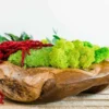 Irregular wooden bowl with light green and dark green moss, red drooping plant, and green leaves on a wooden surface.