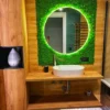 Round mirror with green LED light, set in a green moss wall, above a wooden vanity and white sink.