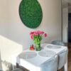 Round green moss wall art in a dark frame mounted on a dining room wall above a table with flowers.