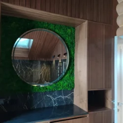 Round mirror mounted on a green moss wall above a black marble counter framed by built-in wooden cabinets.