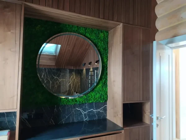 Round mirror mounted on a green moss wall above a black marble counter framed by built-in wooden cabinets.