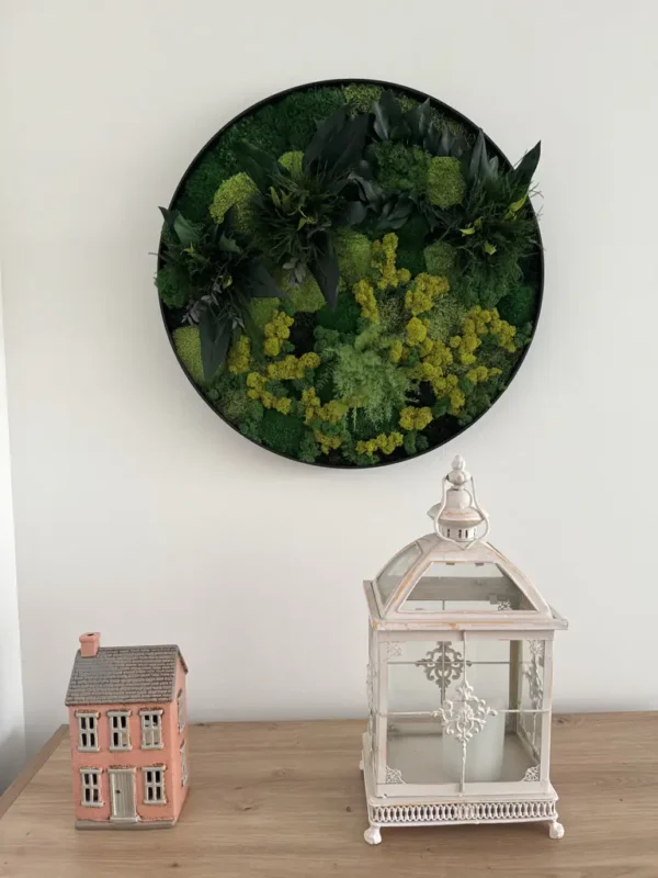 Round green moss wall art above a wooden table holding a ceramic house figurine and a white candle lantern.