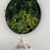 Circular moss wall art in a black frame with green and yellow foliage, hanging above a white lantern.