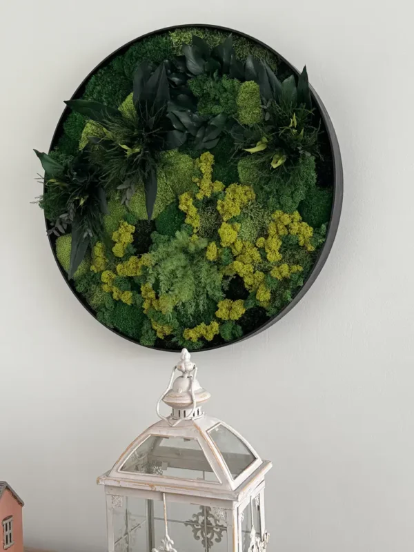 Circular moss wall art in a black frame with green and yellow foliage, hanging above a white lantern.