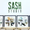Green moss letters spelling SASH STUDIO mounted on a white wall above salon vanity mirrors and chairs.