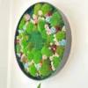 Angled view of round wall art with textured moss and pastel flowers set in deep black frame against white wall