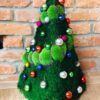 Green moss Christmas tree with colorful mini ornaments and purple bead garland against an exposed brick wall