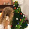 Green moss Christmas tree decorated with colorful ornaments and purple bead garland on a table next to a child.