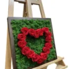 Green moss wall art in a dark wood frame featuring a heart made of red roses, displayed on an easel.
