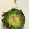 Front view of round wall clock with wood slab dial, preserved green moss, and lush foliage displayed on an easel