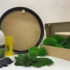 Round DIY moss art kit with black frame, glue, yellow spreader, gloves, and open box packed with green preserved moss.