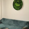 Round preserved moss and fern wall art in white frame hanging on wall above a green sofa.