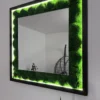 Angled view of a square moss mirror with black wood frame and glowing LED backlighting on a white wall.