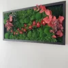 Angled view of rectangular wall art with textured green moss, red flowers, and eucalyptus in a black frame.