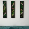 Three vertical black-framed moss wall panels with colorful flower paths hanging above a teal velvet couch.