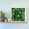 Front view of square moss and pink flower wall art in a white frame, displayed on a wooden shelf next to potted plants.
