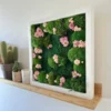 Side view of square moss and pink flower wall art in a white frame, displayed on a wooden shelf beside potted cacti.
