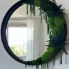 Round mirror with dark frame, partially covered by green moss and fern foliage, hanging on a white wall.
