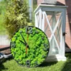 Round wall clock filled with green reindeer moss, black frame, and numerals, resting on grass by a white lantern.