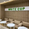 Green moss letters spelling WHITE CUP on a white panel mounted above upholstered seating in a cafe.
