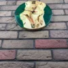 Front view of round maple wood and emerald green resin wall clock with black Roman numerals on a brick wall