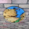Round oak wood wall clock featuring blue and green epoxy resin inlay and black numerals, mounted on a brick wall.