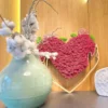 Magenta moss heart wall art framed in wood, standing against wood paneling behind a pale green ceramic vase