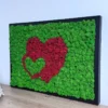 Angled view of black framed moss art with red and green double hearts design on a light wood tabletop.