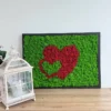 Black framed wall art with bright green moss and two red nested hearts resting on a wooden desk by a lantern.