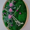 Close-up of round moss wall art with pink roses, eucalyptus, and white branches inside a natural wood frame