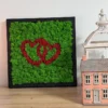 Square black framed green moss wall art with two red hearts on a wooden shelf next to a lantern