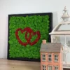 Framed green and red moss heart wall art displayed on a wooden table beside a miniature ceramic house