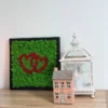 Square moss wall art with red interlocking hearts on green lichen in a black frame, displayed on a wooden shelf