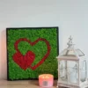 Green moss wall art with red hearts in a black frame, displayed on a wooden table beside a lit candle and white lantern.