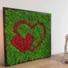 Angled view of green moss wall art with red double hearts in a black frame, displayed on a wood floor against a white wall.