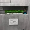 Rectangular glass planter with green moss and white stones on a gray tiled bathroom shelf above a flush plate.