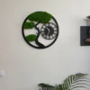Round black tree wall clock with green reindeer moss foliage, mounted on a white wall above a palm plant