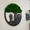 Black tree of life wall clock with green bun moss canopy and couple silhouette, hanging on a white wall.