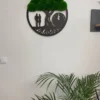 Round tree of life wall clock with green bun moss and black couple silhouette mounted on a white wall above a plant.