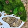 Round tree of life wall art with green moss and pinecones in a white cutout frame over tree bark background.