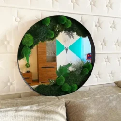 Black-framed round mirror with green moss and plants, mounted on a white tufted wall above a beige sofa.