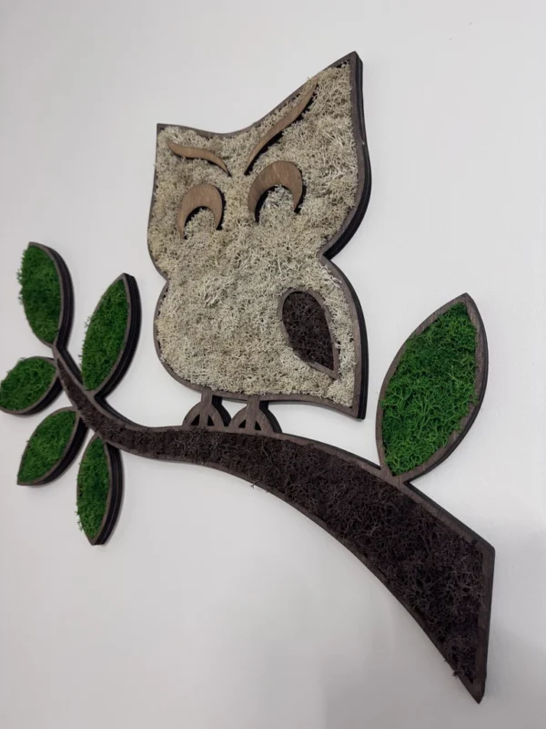 Moss art in owl shape on a branch with green leaves and light moss plumage on a white wall.