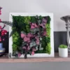 IMG_8677_result Square plant wall art with green reindeer moss, fern, and red eucalyptus leaves in a white frame on a wooden shelf.