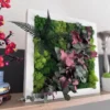 IMG_8679_result Square plant wall art with green reindeer moss, red eucalyptus, and dark green fern leaves in a white wooden frame on a wooden shelf.