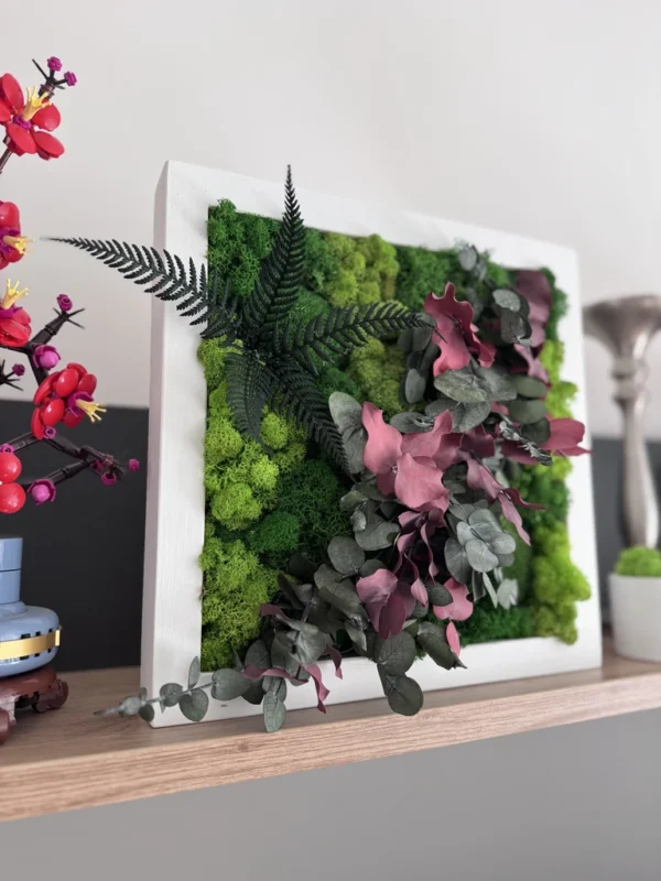 IMG_8679_result Square plant wall art with green reindeer moss, red eucalyptus, and dark green fern leaves in a white wooden frame on a wooden shelf.